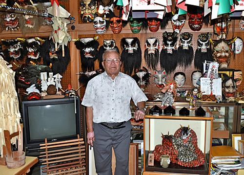私設の神楽殿に立つ竹内惟臣さん。石見神楽面のコレクションが壁一面に並ぶ