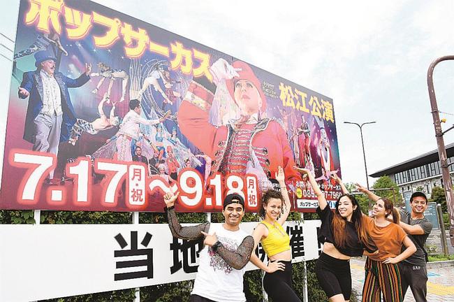 会場予定地に到着し、看板の前でポーズを決めるパフォーマーら＝松江市学園南１丁目