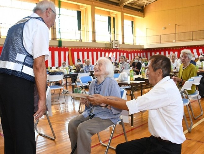 「１００歳をめざす集い」で記念品を受け取る女性（中央）＝９月１６日、浜田市長浜町の長浜小学校体育館