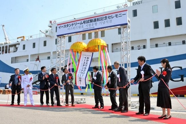試験運航の開始を記念し、くす玉を割って祝うトゥウォン商船の李錫基社長（左から５人目）、平井伸治知事（同６人目）ら＝境港市竹内団地