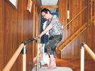 訪問リハビリテーションを受けられず、自宅で手すりにつかまり一人で足の運動をする木村槙江さん＝出雲市大社町宇龍