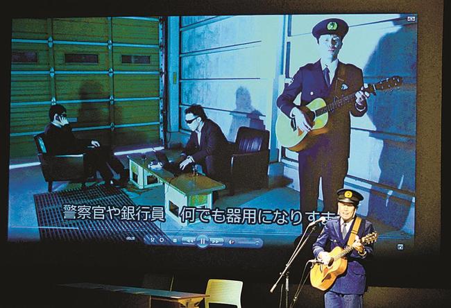 動画を映すスクリーンの前で熱唱する加藤聖規巡査部長＝安来市飯島町、市総合文化ホール