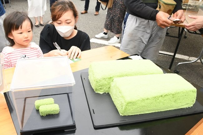 巨大な若草の重さを推測する親子連れ。左は通常サイズ＝松江市八束町波入、彩雲堂八束工場