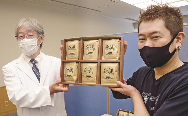 贈呈式でパッケージに「感謝」と書いたコーヒーを掲げる小阪真二病院長（左）と門脇裕二代表＝出雲市姫原４丁目、島根県立中央病院
