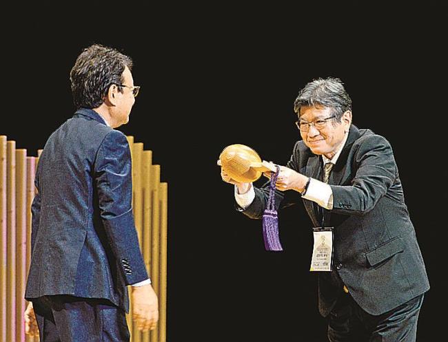 松尾倫男会長（左）から打ち出の小づちを受け取る沖縄大会組織委員会の玻名城泰山副会長＝松江市殿町、島根県民会館