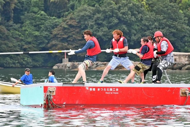 海上の台船の上で、力いっぱい綱を引く参加者＝松江市美保関町北浦、北浦海水浴場