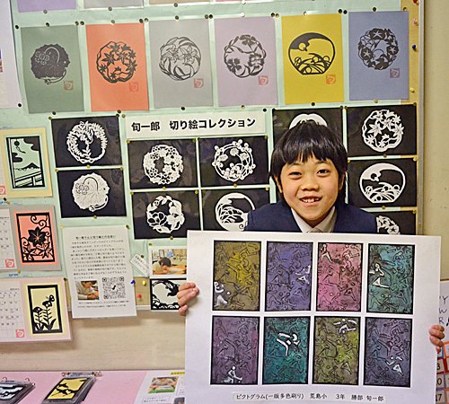 切り絵作品を背に絵画を見せる勝部旬一郎君。手にするのはピクトグラムの版画＝安来市荒島町、荒島小学校