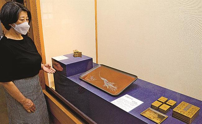 角度によって色が異なって見える「鮎蒔絵香盆」（中央）の説明をする佐々木杏里学芸員＝出雲市大社町杵築西、手銭美術館