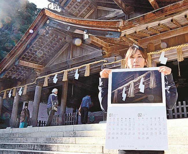 美保神社を背景にカレンダーを手にする松江観光協会美保関町支部の職員＝松江市美保関町美保関