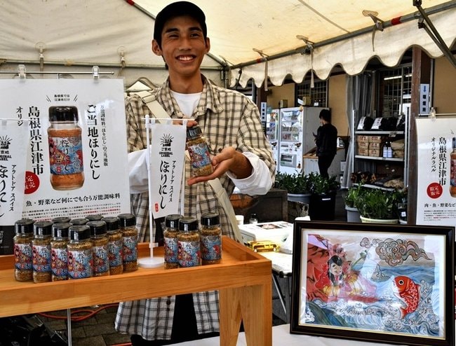 タイを釣り上げる「恵比寿」の絵がラベルに採用された商品をＰＲする佐々木和仁さん（左）＝江津市後地町、道の駅「サンピコごうつ」