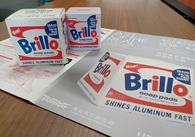 「ブリロの箱」を模した小箱に入ったお菓子（左）と、ブリロの箱が表紙に印刷された開館記念展のリーフレット