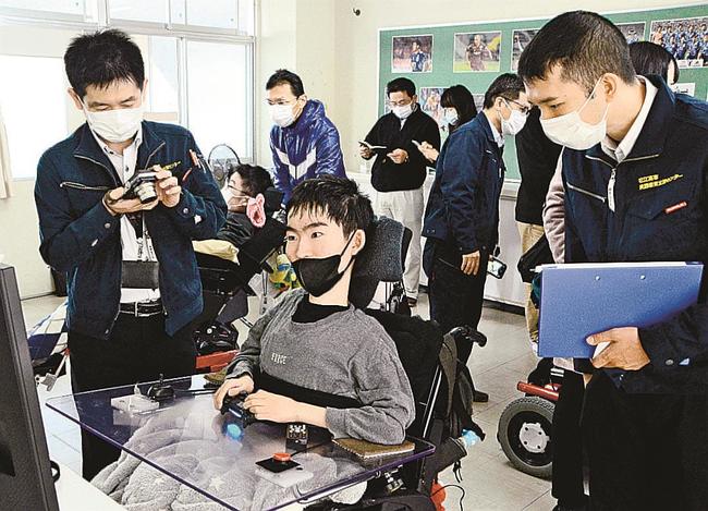 生徒（中央）が握るコントローラーを視察する松江工業高等専門学校の教員＝松江市上乃木５丁目、松江緑が丘養護学校