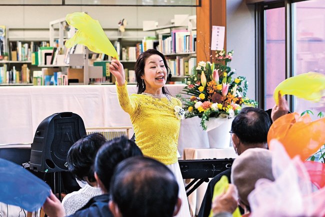 スカーフを振りながら歌声を披露する田部由美子さん＝安来市安来町、市立図書館