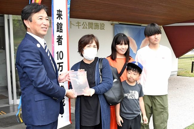 １０万人目の来館者になり、飯塚俊之市長（左）から記念品を受け取る田辺千恵子さん（左から２人目）ら＝出雲市西新町２丁目、市トキ公開施設