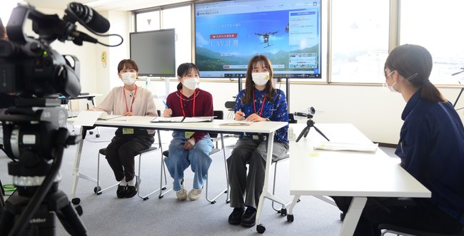 動画撮影用のカメラ（左）を設置し共立エンジニヤの女性社員（右）を取材する学生＝松江市西津田２丁目