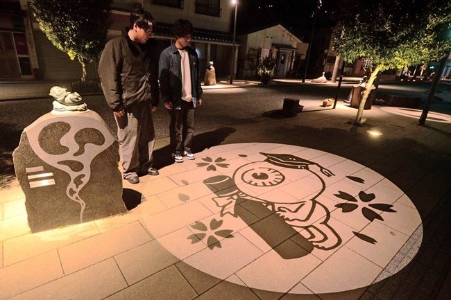 帽子をかぶり、賞状筒を手にした目玉おやじの妖怪影絵に見入る観光客＝境港市大正町