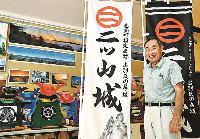 二ツ山城の旗や、イベントで子どもが着用する段ボール甲冑を前に来場を呼びかける吉川正さん＝島根県邑南町下田所、田所公民館