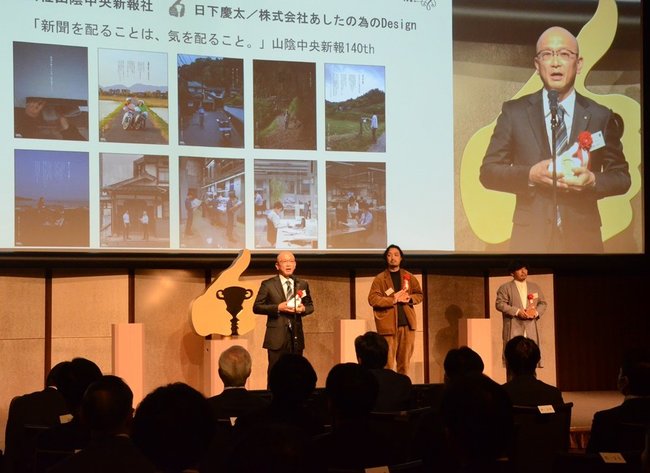 トロフィーを胸に受賞あいさつをする山陰中央新報社の大庭浩史取締役兼読者局長(壇上左)=東京都千代田区、東京会館