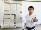 人工ルビー合成の実験過程を英語で書いたポスターの横で、空手の形を披露する坂本來愛さん＝松江市西津田９丁目、開星高校