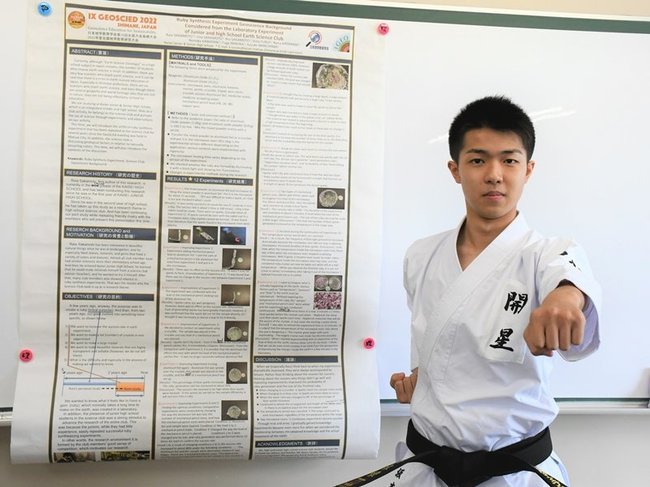 人工ルビー合成の実験過程を英語で書いたポスターの横で、空手の形を披露する坂本來愛さん＝松江市西津田９丁目、開星高校