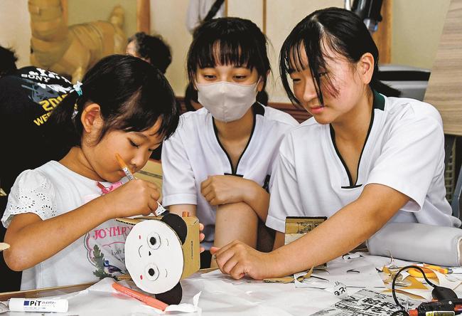 ボランティアの安来一中生と段ボール貯金箱を作る児童（左）＝安来市上坂田町、赤江交流センター