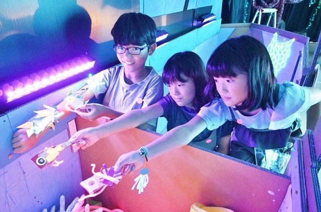 自作の魚飾りをライトに照らす子どもたち＝浜田市野原町、市世界こども美術館